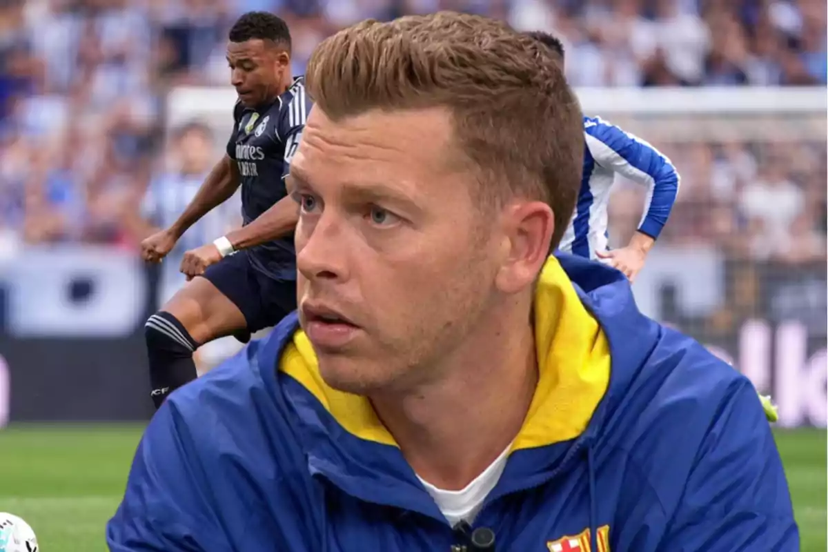 A man in a blue and yellow jacket appears in the foreground while a soccer match is being played in the background between two teams wearing dark and light uniforms.