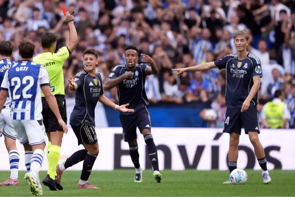 A referee shows a red card during a soccer match while the players protest and the crowd watches from the stands. A referee shows a red card during a soccer match while the players protest and the crowd watches from the stands.