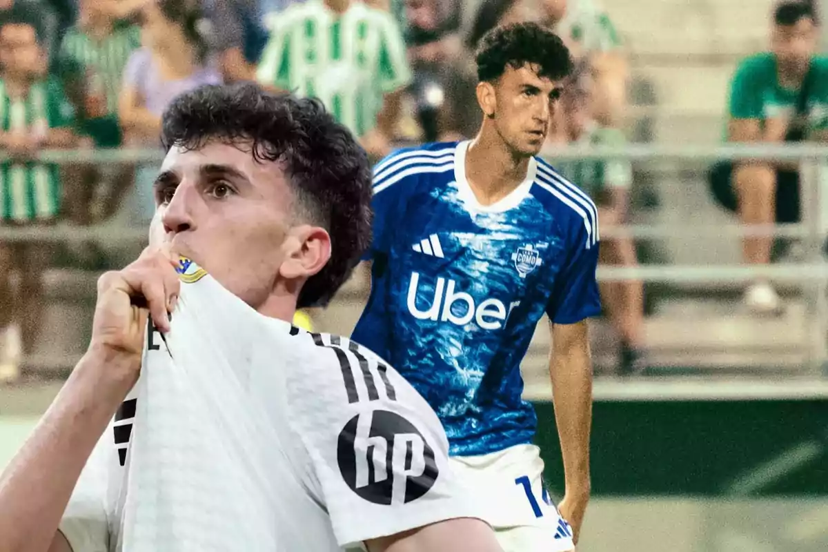 A player kisses the crest on his white jersey while another player in a blue uniform is in the background at a stadium with a blurred crowd.