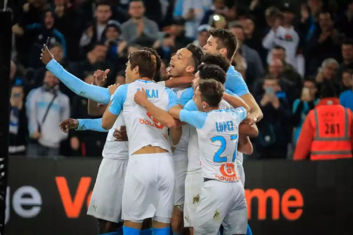 Jugadores de fútbol celebran un gol abrazándose en el campo mientras la afición los observa emocionada desde las gradas Jugadores de fútbol celebran un gol abrazándose en el campo mientras la afición los observa emocionada desde las gradas