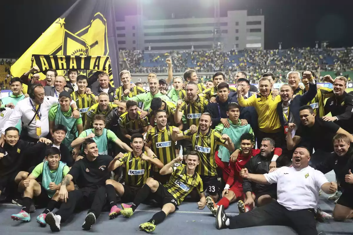 Players and coaching staff of a soccer team celebrate together on the field with a large yellow and black flag and a scarf that says Airat Almaty, while the fans watch them from the stands. Players and coaching staff of a soccer team celebrate together on the field with a large yellow and black flag and a scarf that says Airat Almaty, while the fans watch them from the stands.