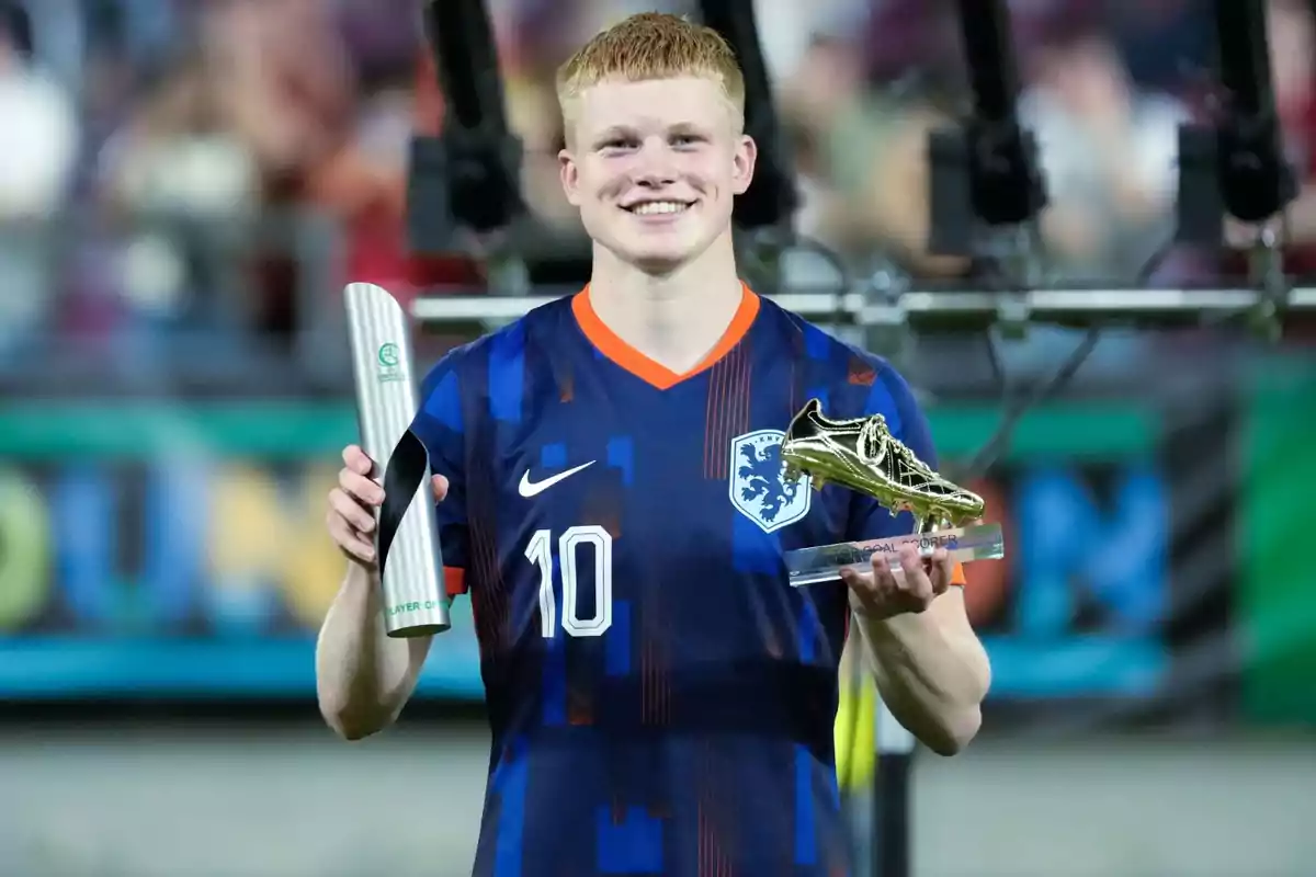 Futbolista con uniforme azul de Países Bajos sonríe mientras sostiene dos trofeos uno en cada mano en un estadio Futbolista con uniforme azul de Países Bajos sonríe mientras sostiene dos trofeos uno en cada mano en un estadio