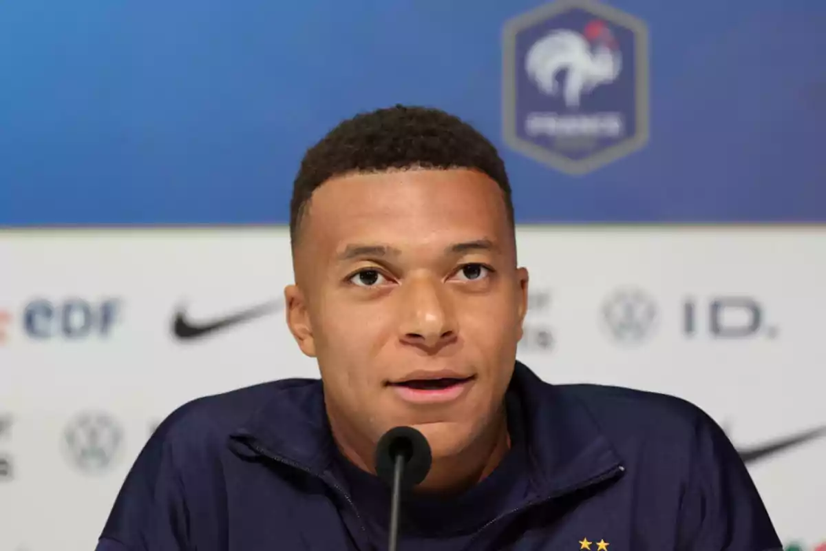 A player in a navy blue jacket speaks in front of a microphone at a press conference with logos and a French crest in the background.
