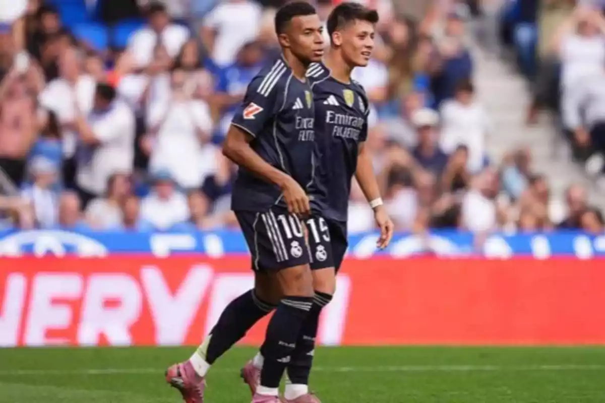 Two players in dark uniforms walk together on the field during a match with spectators in the stands in the background.