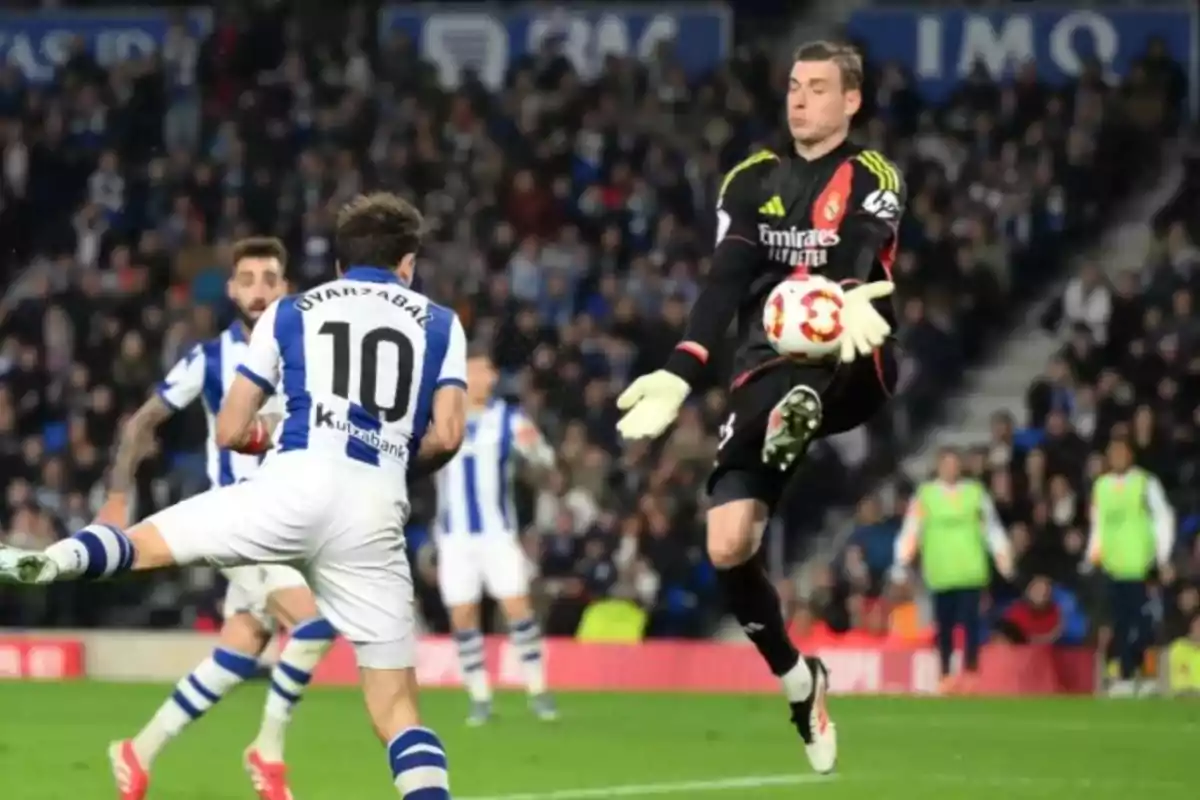 Goalkeeper dressed in black stopping a ball in front of a player wearing a white and blue uniform during a soccer match in a stadium full of spectators Goalkeeper dressed in black stopping a ball in front of a player wearing a white and blue uniform during a soccer match in a stadium full of spectators