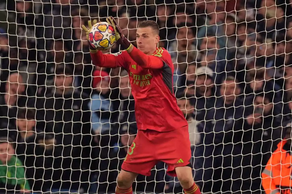 Soccer goalkeeper in a red uniform saving a ball in front of the goal during a match with a crowd in the background Soccer goalkeeper in a red uniform saving a ball in front of the goal during a match with a crowd in the background