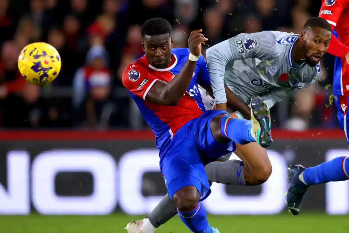 Two players compete for the ball during a match, one wearing a blue and red uniform and the other in gray, both showing intensity in the play. Two players compete for the ball during a match, one wearing a blue and red uniform and the other in gray, both showing intensity in the play.