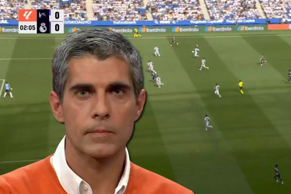 A gray-haired man in an orange sweater appears in the foreground with a soccer match in the background at a stadium full of spectators.