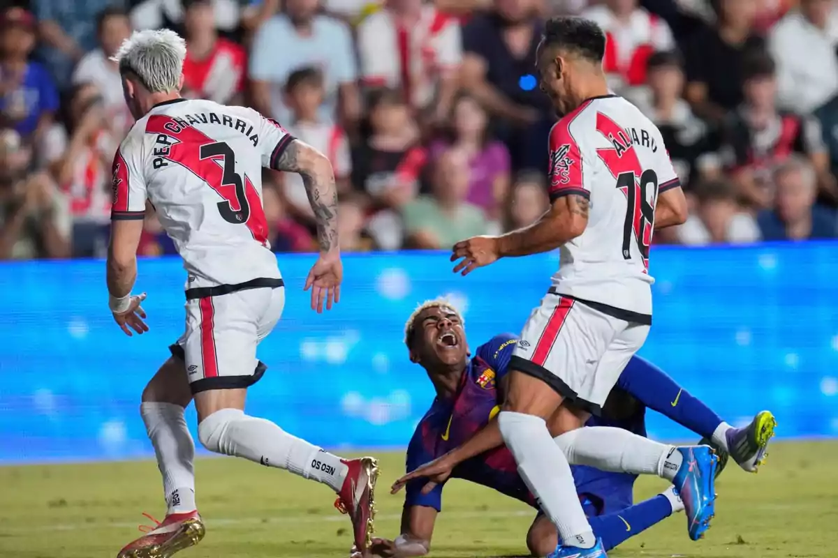 Two players in white uniforms and one in a blue uniform fight for the ball as the player in blue falls to the ground with a look of pain during a match. Two players in white uniforms and one in a blue uniform fight for the ball as the player in blue falls to the ground with a look of pain during a match.