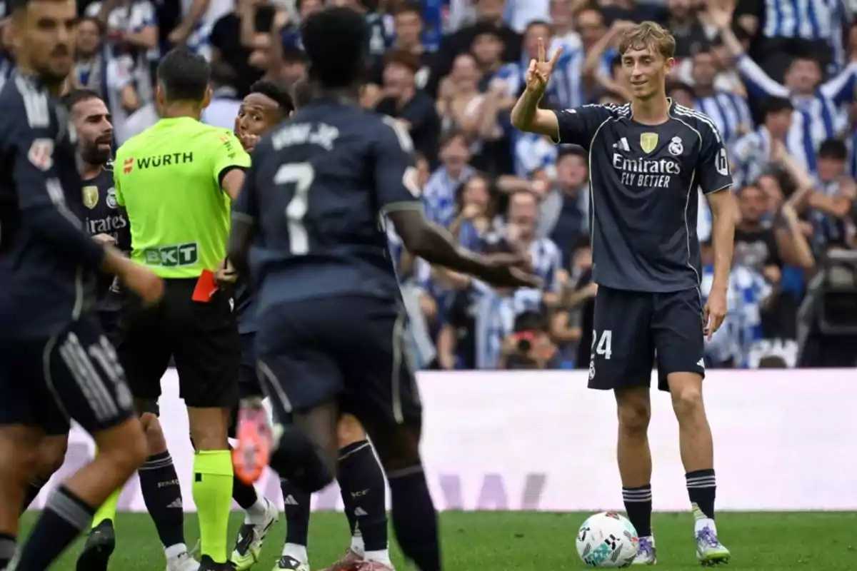 Un jugador de fútbol del Real Madrid sonríe y hace una señal de victoria con los dedos mientras otros jugadores y el árbitro discuten en el fondo durante un partido Un jugador de fútbol del Real Madrid sonríe y hace una señal de victoria con los dedos mientras otros jugadores y el árbitro discuten en el fondo durante un partido