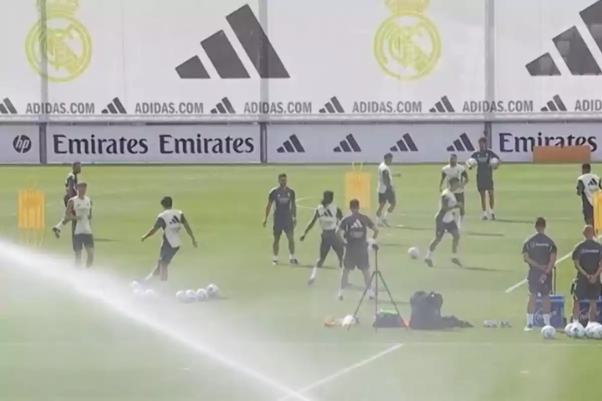 Players and coaches of a soccer team train on a field with sprinklers on and several balls, while in the background there are sponsor advertisements and a club crest. Players and coaches of a soccer team train on a field with sprinklers on and several balls, while in the background there are sponsor advertisements and a club crest.