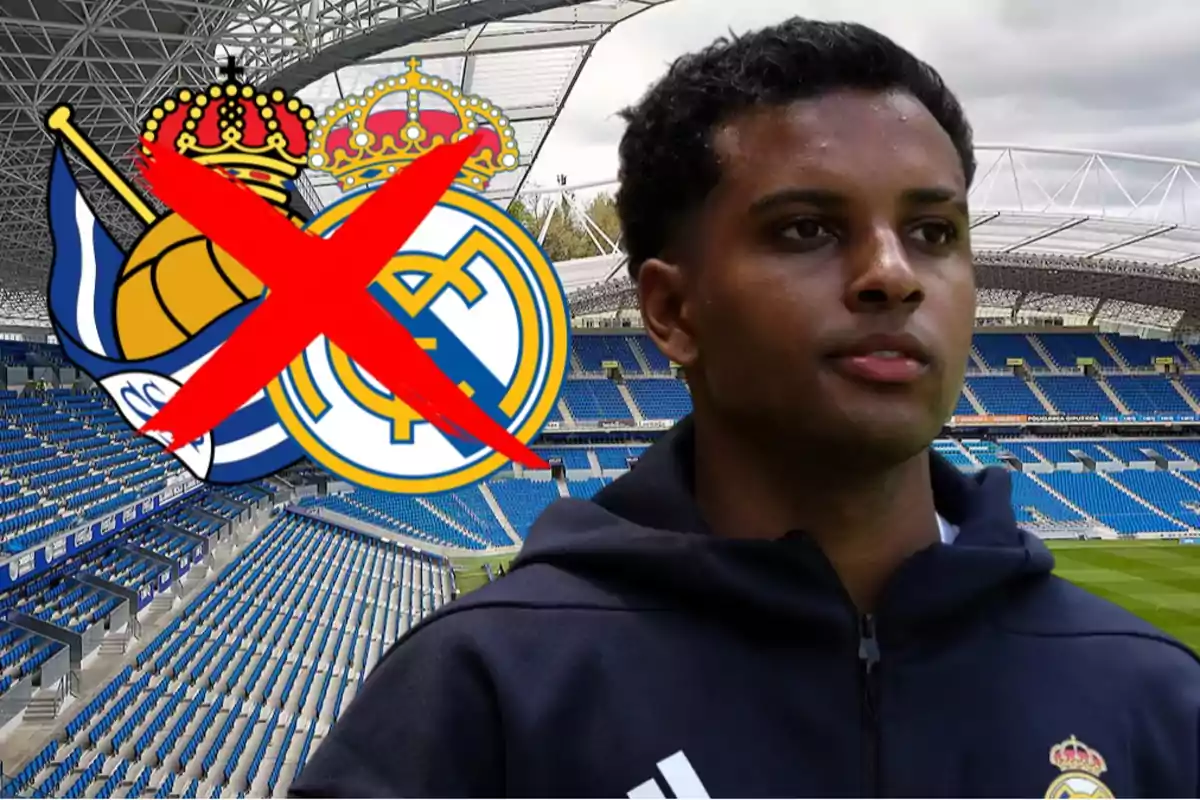 Un futbolista con chaqueta del Real Madrid en un estadio vacío con los escudos de la Real Sociedad y el Real Madrid al fondo y una gran X roja sobre ellos
