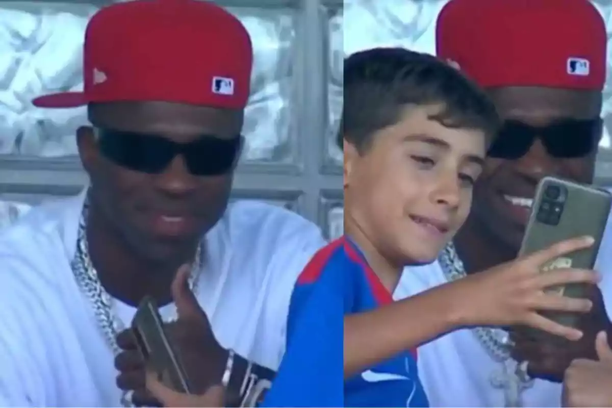 A man wearing a red cap and dark glasses smiles as he poses next to a boy taking a selfie with his mobile phone. A man wearing a red cap and dark glasses smiles as he poses next to a boy taking a selfie with his mobile phone.