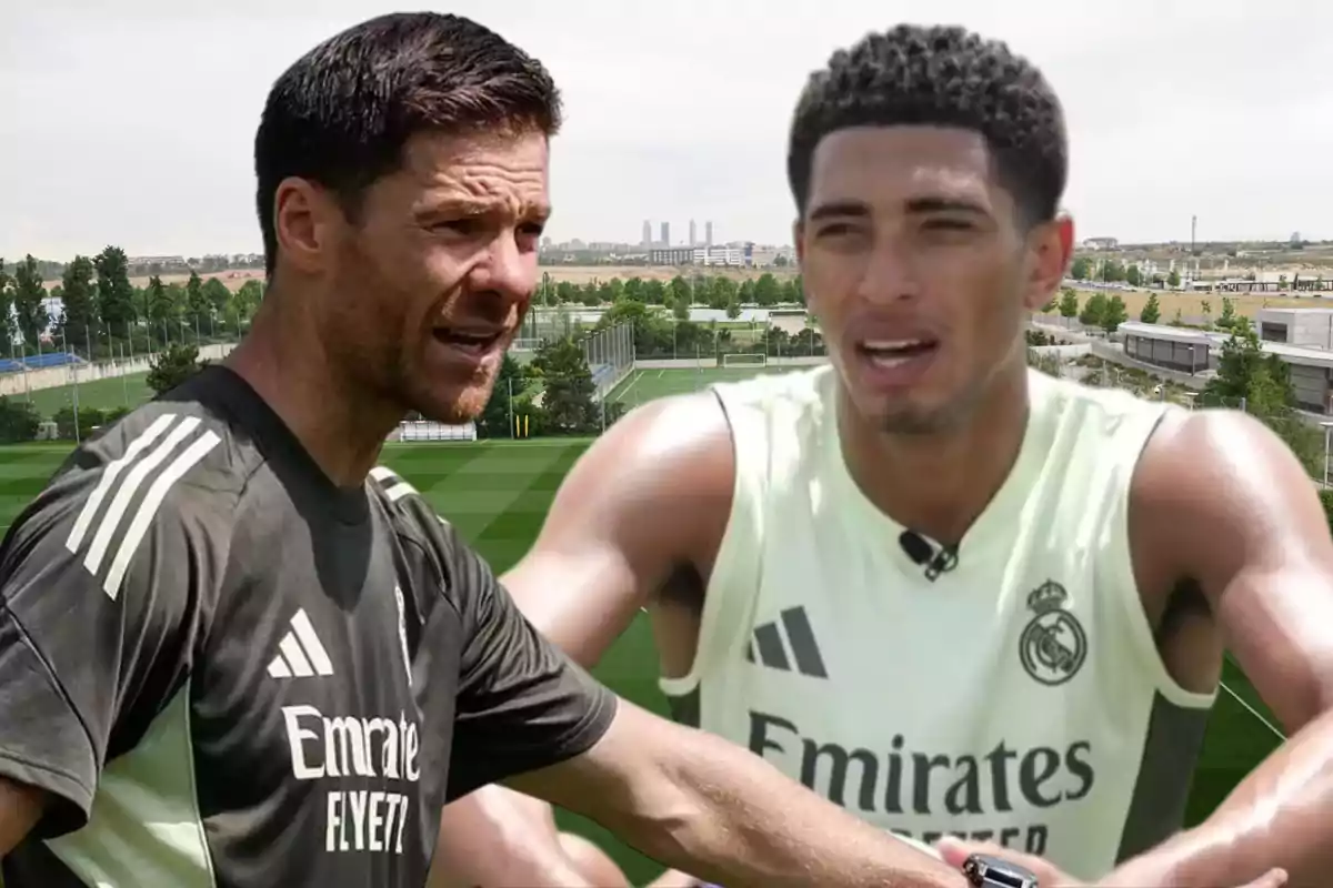 Two Real Madrid players talk on a training field with grass and buildings in the background.