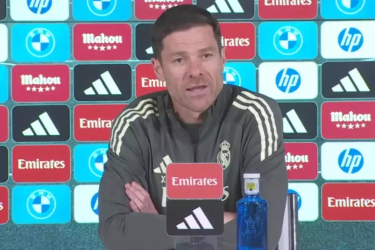 Entrenador de fútbol con chaqueta del Real Madrid hablando en una rueda de prensa frente a un fondo con logotipos de patrocinadores