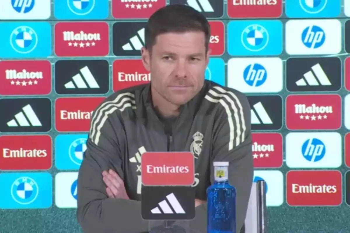 Entrenador de fútbol del Real Madrid sentado en una rueda de prensa con fondo de patrocinadores y una botella de agua sobre la mesa