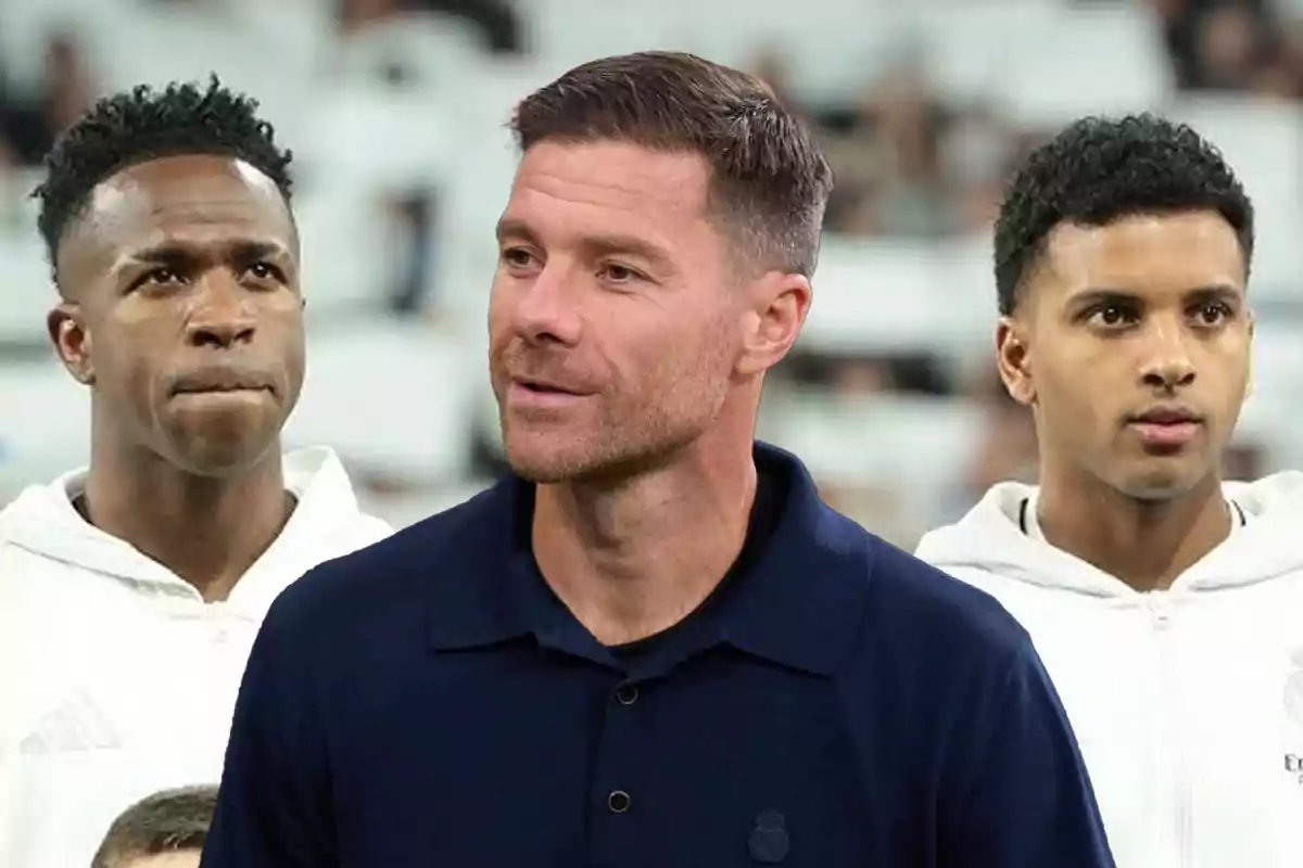 Tres hombres con expresión seria en un estadio, dos de ellos con chaquetas blancas y uno al centro con camisa azul oscuro
