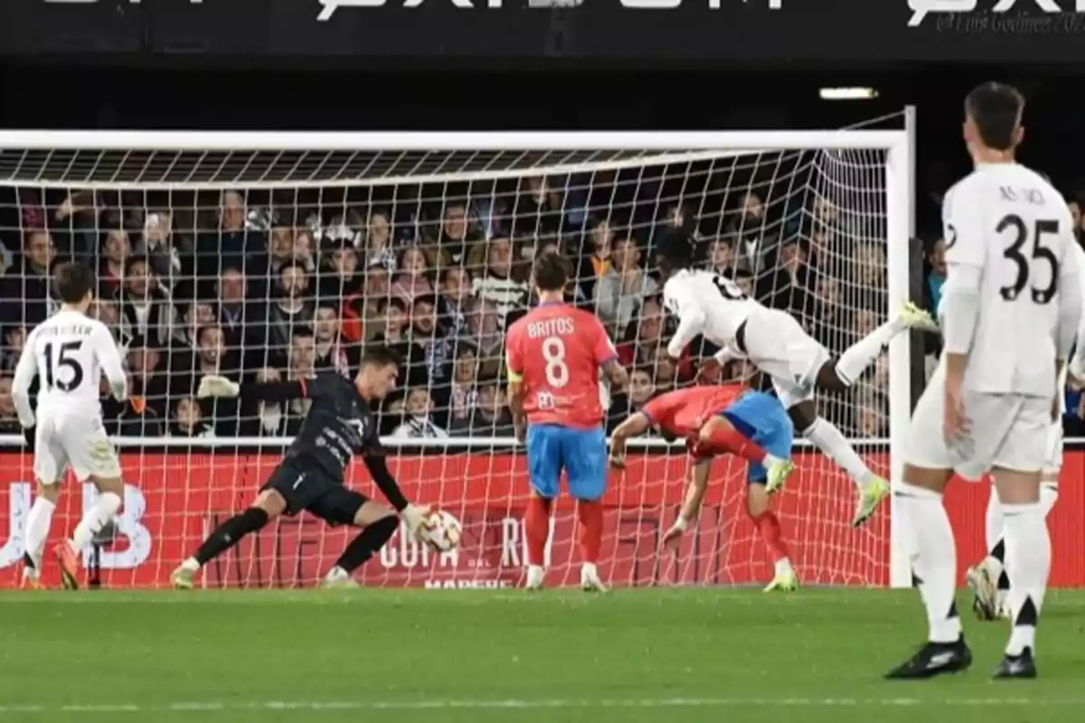 A soccer player in a white uniform jumps to head the ball toward the goal as the goalkeeper and other players watch the action in a stadium full of spectators. A soccer player in a white uniform jumps to head the ball toward the goal as the goalkeeper and other players watch the action in a stadium full of spectators.