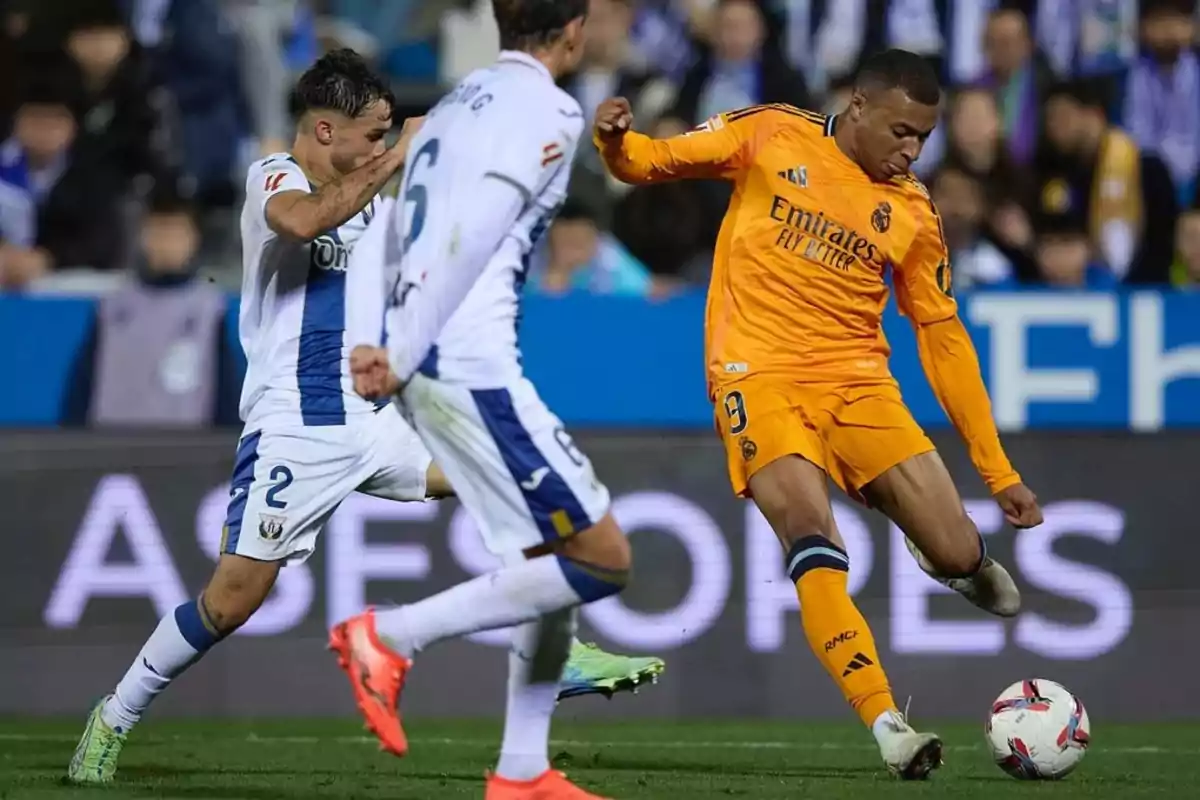 Jugadores de fútbol en acción durante un partido, uno de ellos con uniforme naranja controlando el balón mientras los otros dos con uniforme blanco intentan defender.