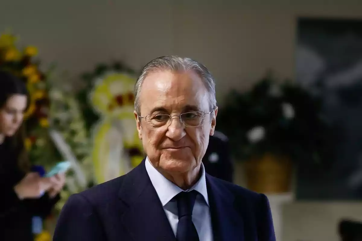 An older man with glasses and a dark suit in an indoor setting with flowers and people in the background. An older man with glasses and a dark suit in an indoor setting with flowers and people in the background.