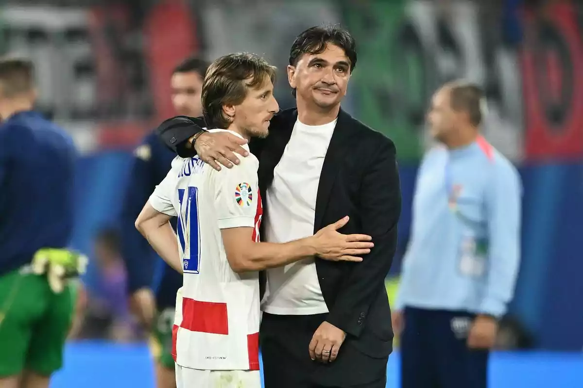 A player in a white and red uniform hugs a man in a black suit on a soccer field.