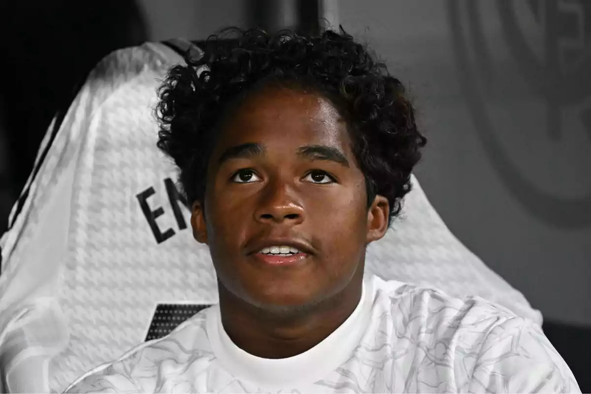 Un joven con camiseta blanca sentado frente a un fondo oscuro.