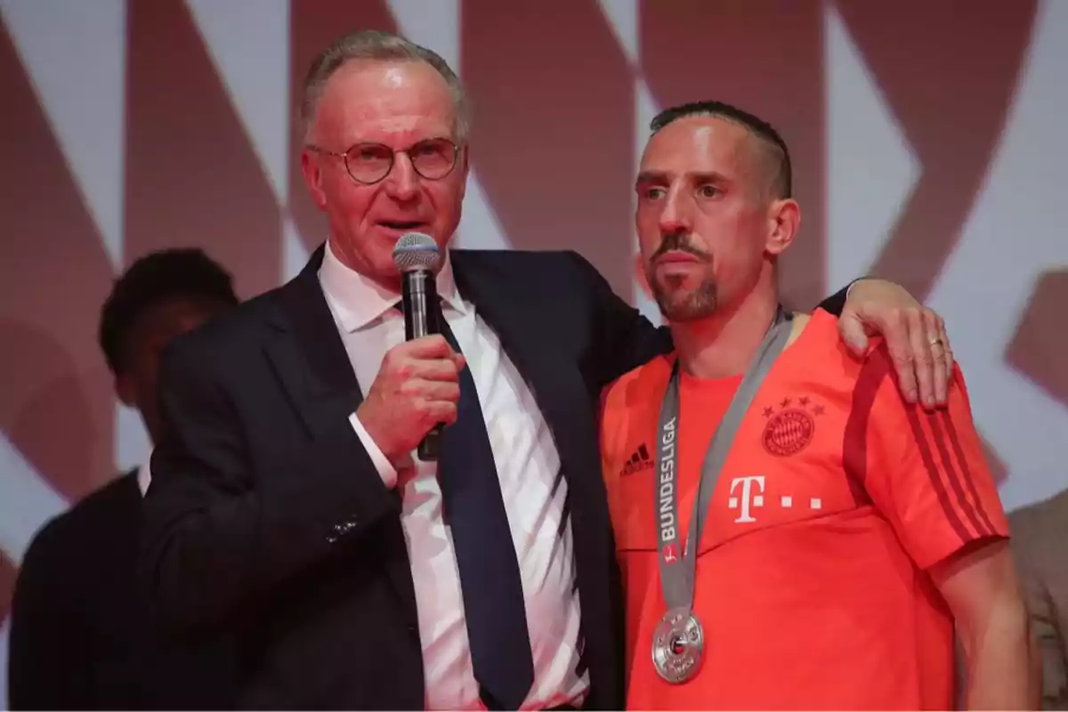 Two men on a stage, one with a suit and microphone, the other with a sports jersey and medal.