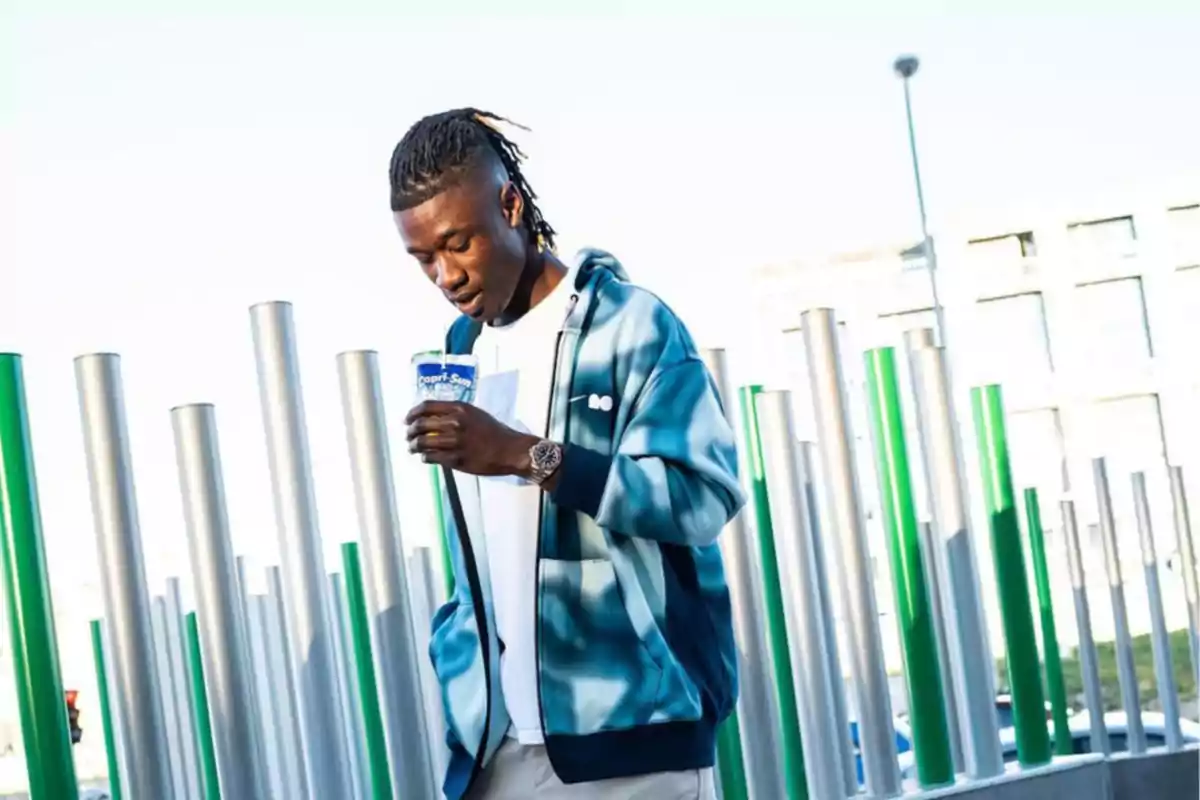 A man in a blue and white jacket holds a drink while walking outdoors surrounded by vertical metal poles. A man in a blue and white jacket holds a drink while walking outdoors surrounded by vertical metal poles.