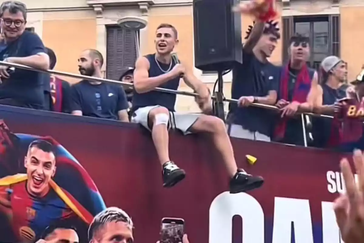 A group of people celebrates on an open-top bus decorated with images of players, one of them is sitting on the edge holding a microphone while others wear team scarves and jerseys.
