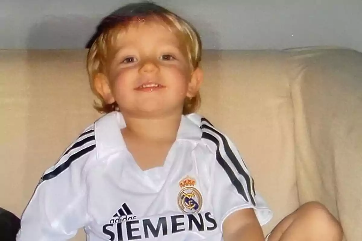Niño pequeño sonriente sentado en un sofá vistiendo una camiseta blanca del Real Madrid