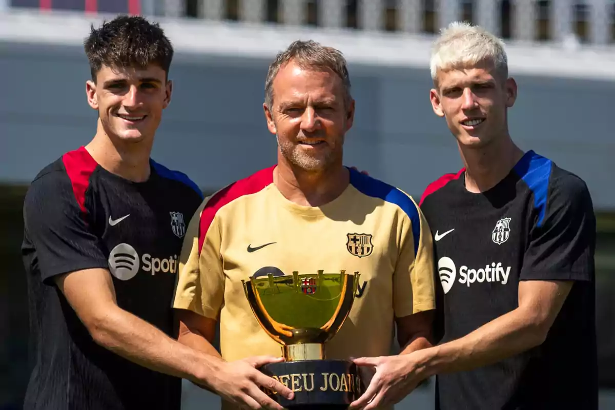 Three people pose with a soccer trophy, two of them wearing black jerseys with red and blue details, and the person in the center wearing a gold jersey. Three people pose with a soccer trophy, two of them wearing black jerseys with red and blue details, and the person in the center wearing a gold jersey.