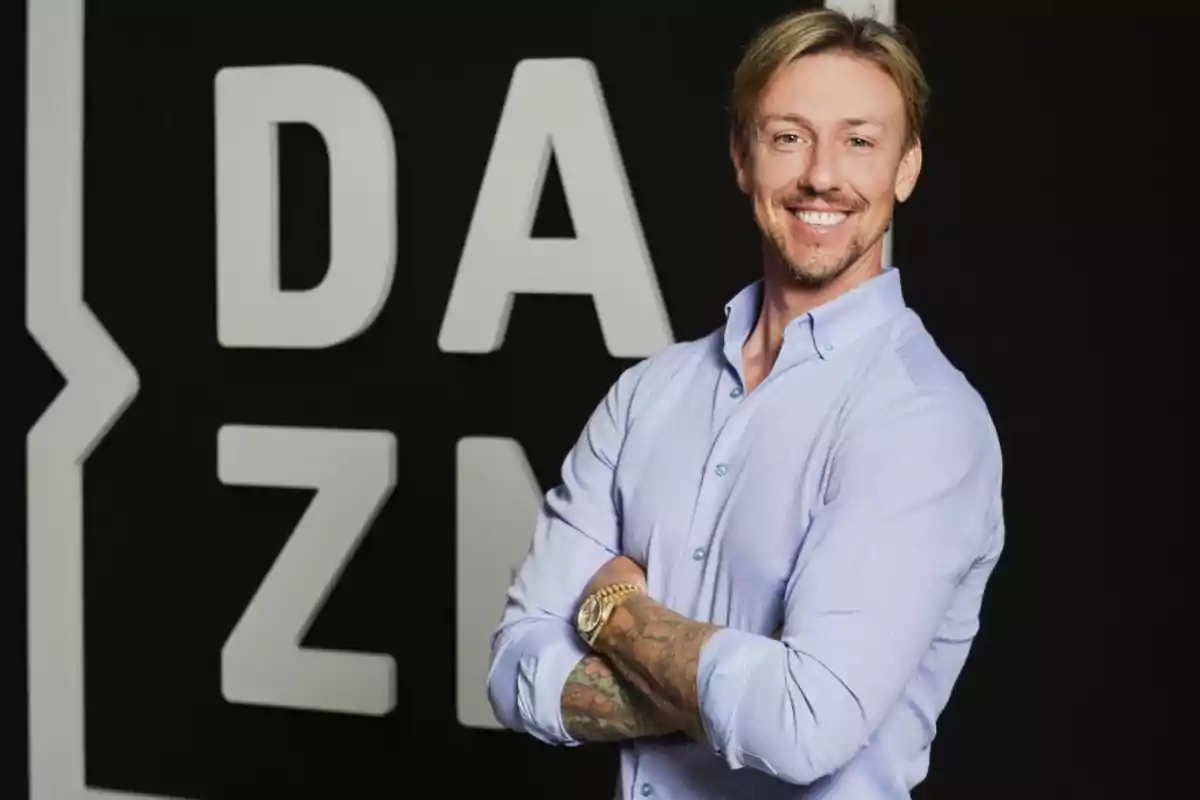 Smiling blond man in a light blue jersey posing with his arms crossed in front of a background with the DAZN logo