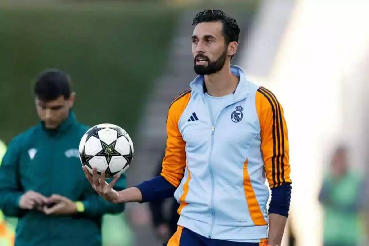 A man in a Real Madrid sports uniform holds a soccer ball in one hand while standing on a field. A man in a Real Madrid sports uniform holds a soccer ball in one hand while standing on a field.