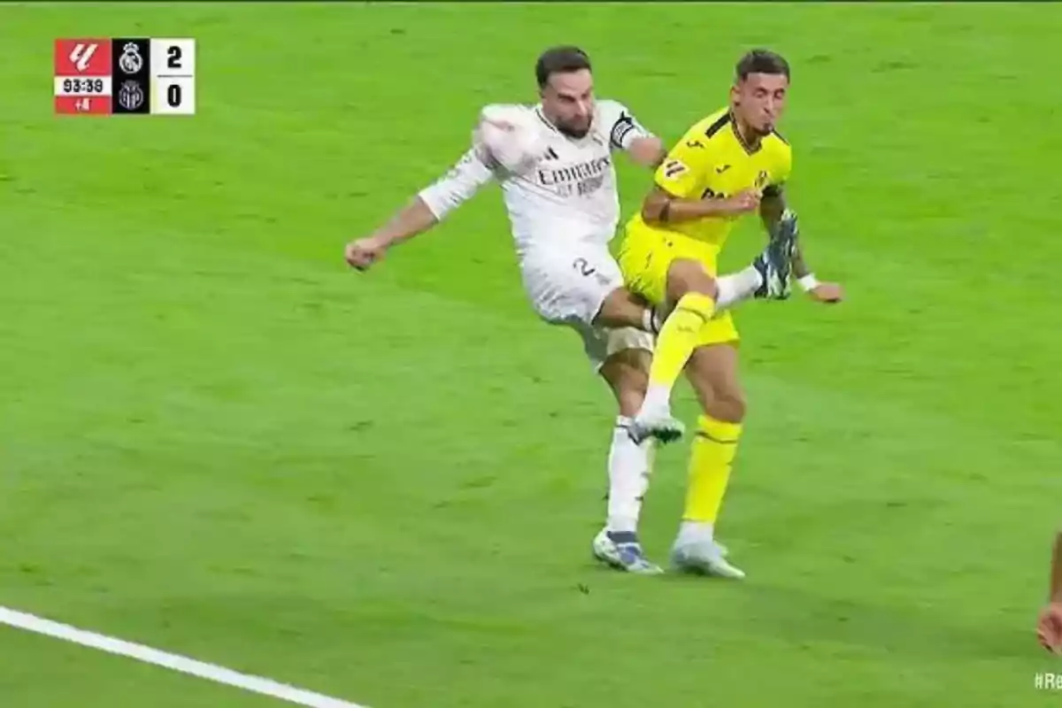 Dos jugadores de fútbol compiten por el balón en un partido, uno vestido de blanco y el otro de amarillo, mientras el marcador muestra 2-0 a favor del equipo de blanco.