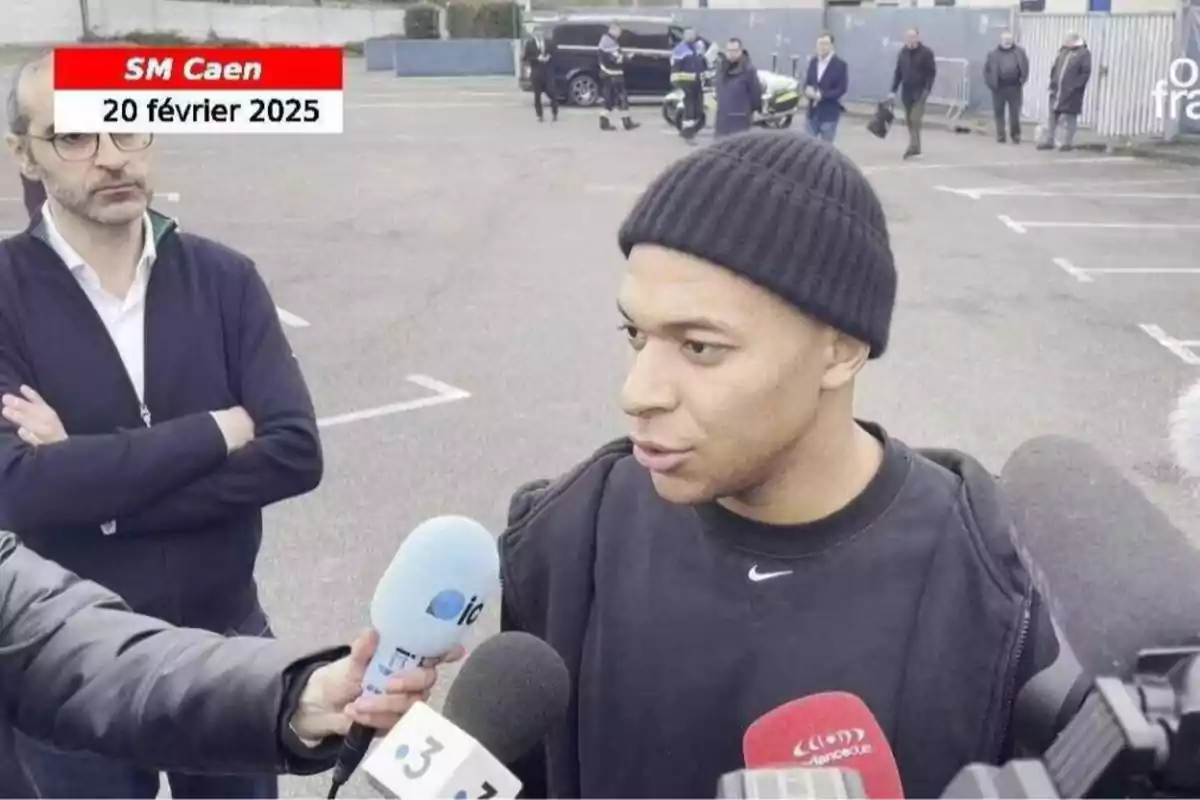 Un hombre con gorro negro es entrevistado por varios medios de comunicación en un estacionamiento, mientras otra persona observa con los brazos cruzados. Un hombre con gorro negro es entrevistado por varios medios de comunicación en un estacionamiento, mientras otra persona observa con los brazos cruzados.