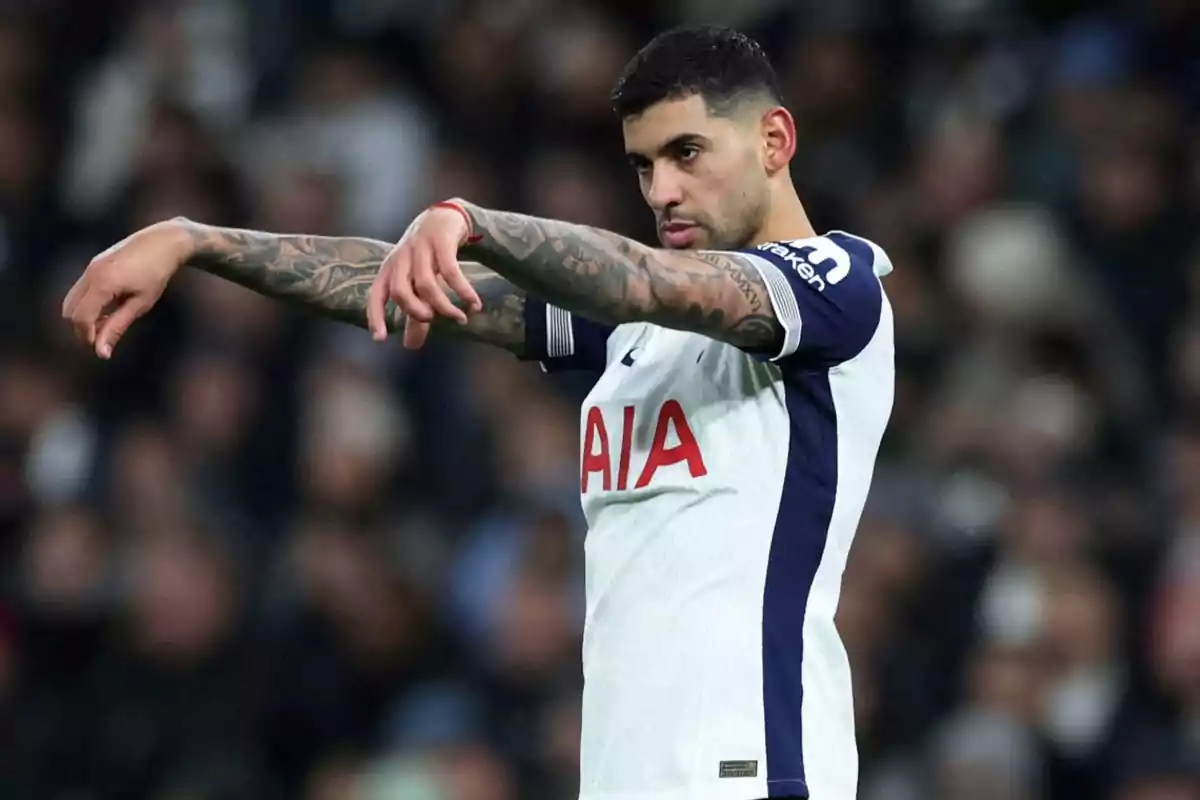 Jugador de fútbol con uniforme blanco y azul en el campo, con los brazos extendidos y tatuajes visibles.