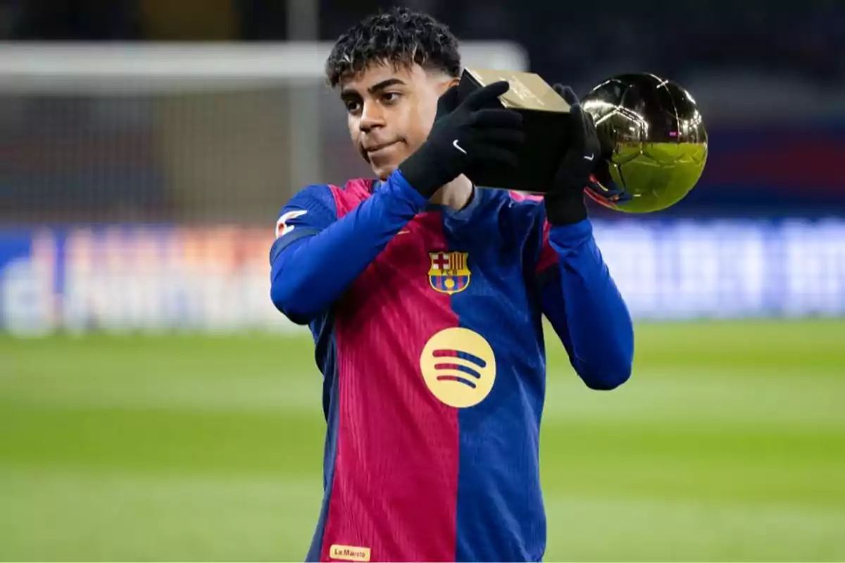 Jugador de fútbol con uniforme azul y rojo sosteniendo un trofeo dorado en un estadio.