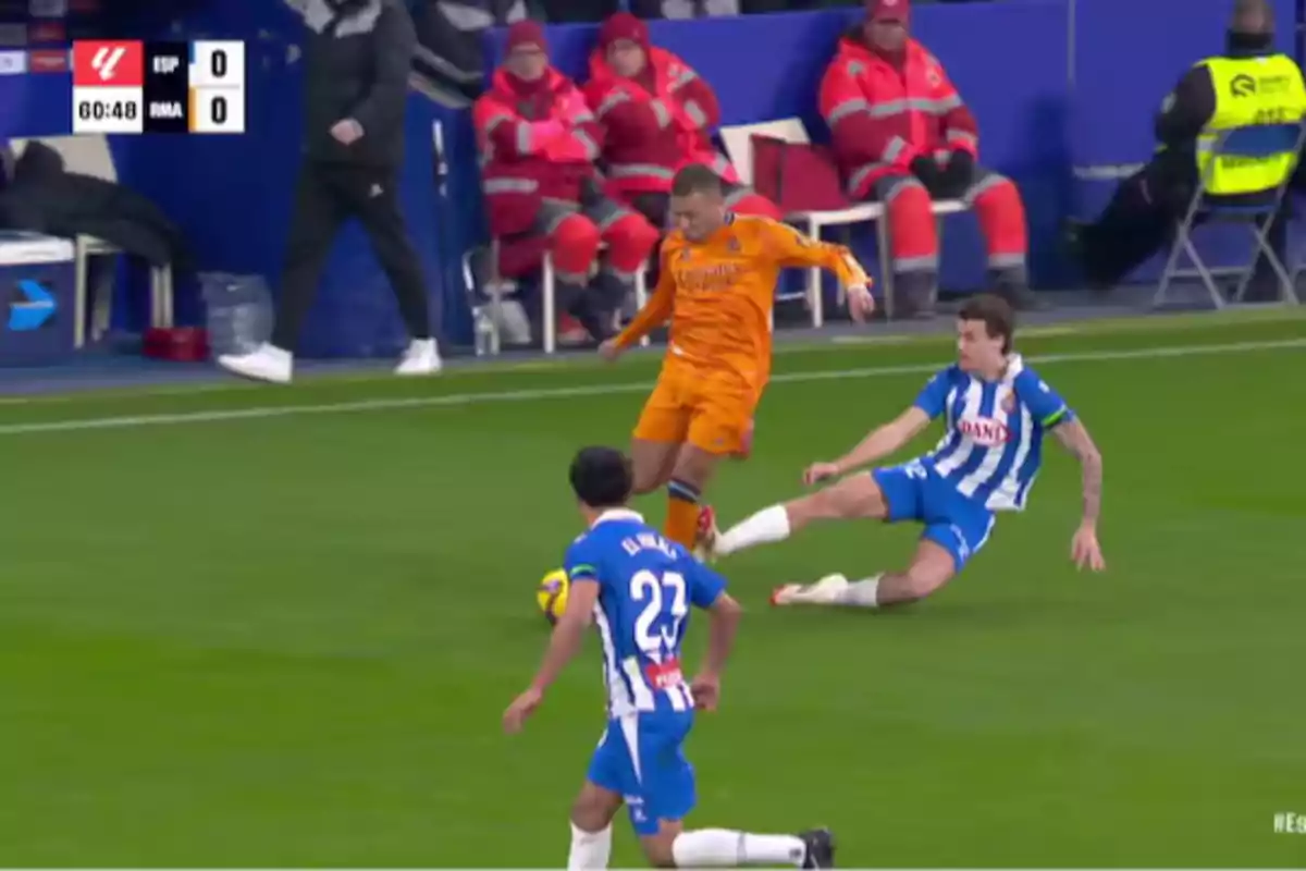 Un jugador de fútbol con uniforme naranja intenta esquivar una entrada de un jugador con uniforme azul y blanco durante un partido, mientras el marcador muestra un empate a cero.