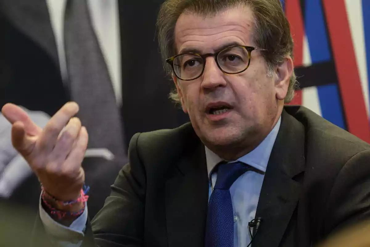 A man with glasses and a dark suit gestures as he speaks. A man with glasses and a dark suit gestures as he speaks.