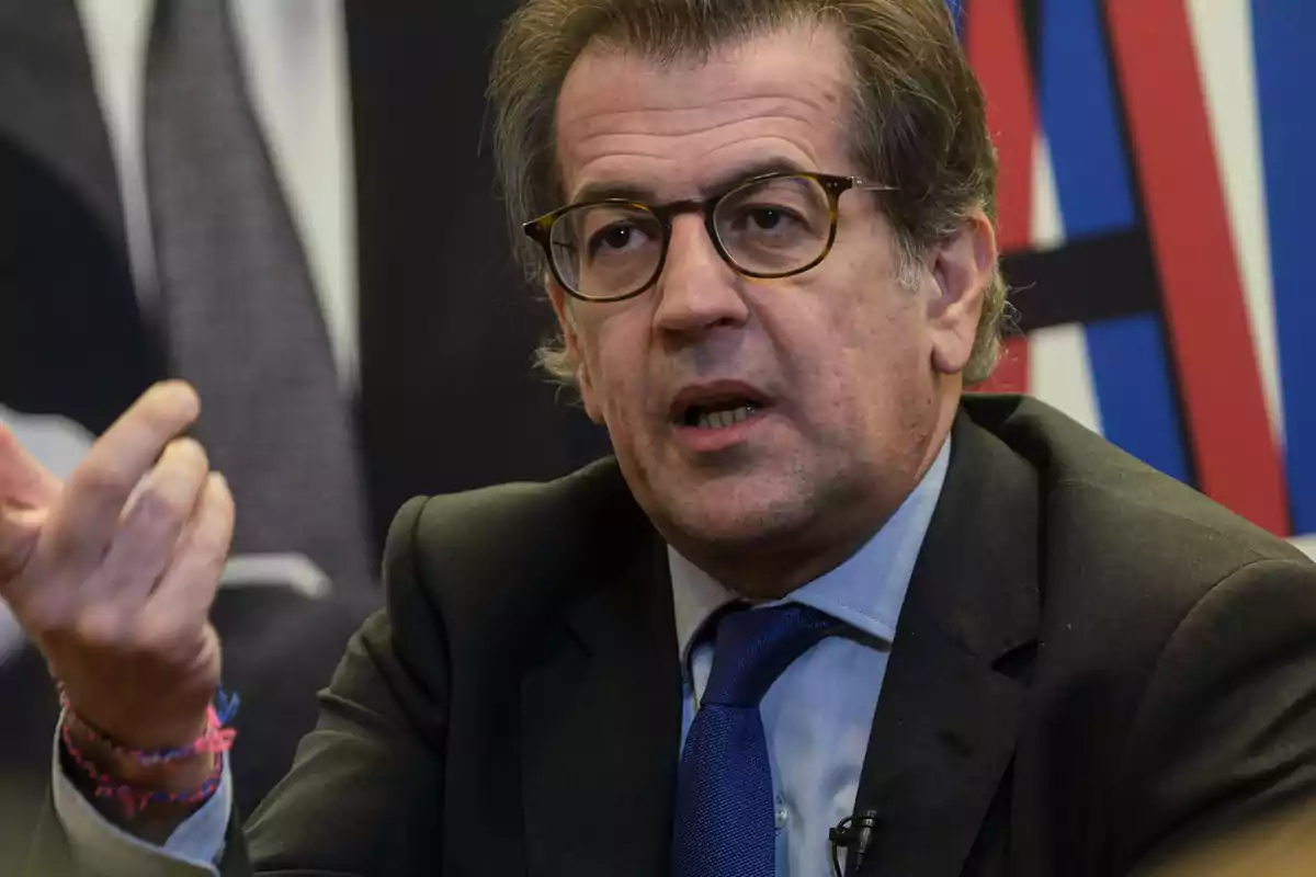 A man with glasses and a dark suit gestures as he speaks. A man with glasses and a dark suit gestures as he speaks.