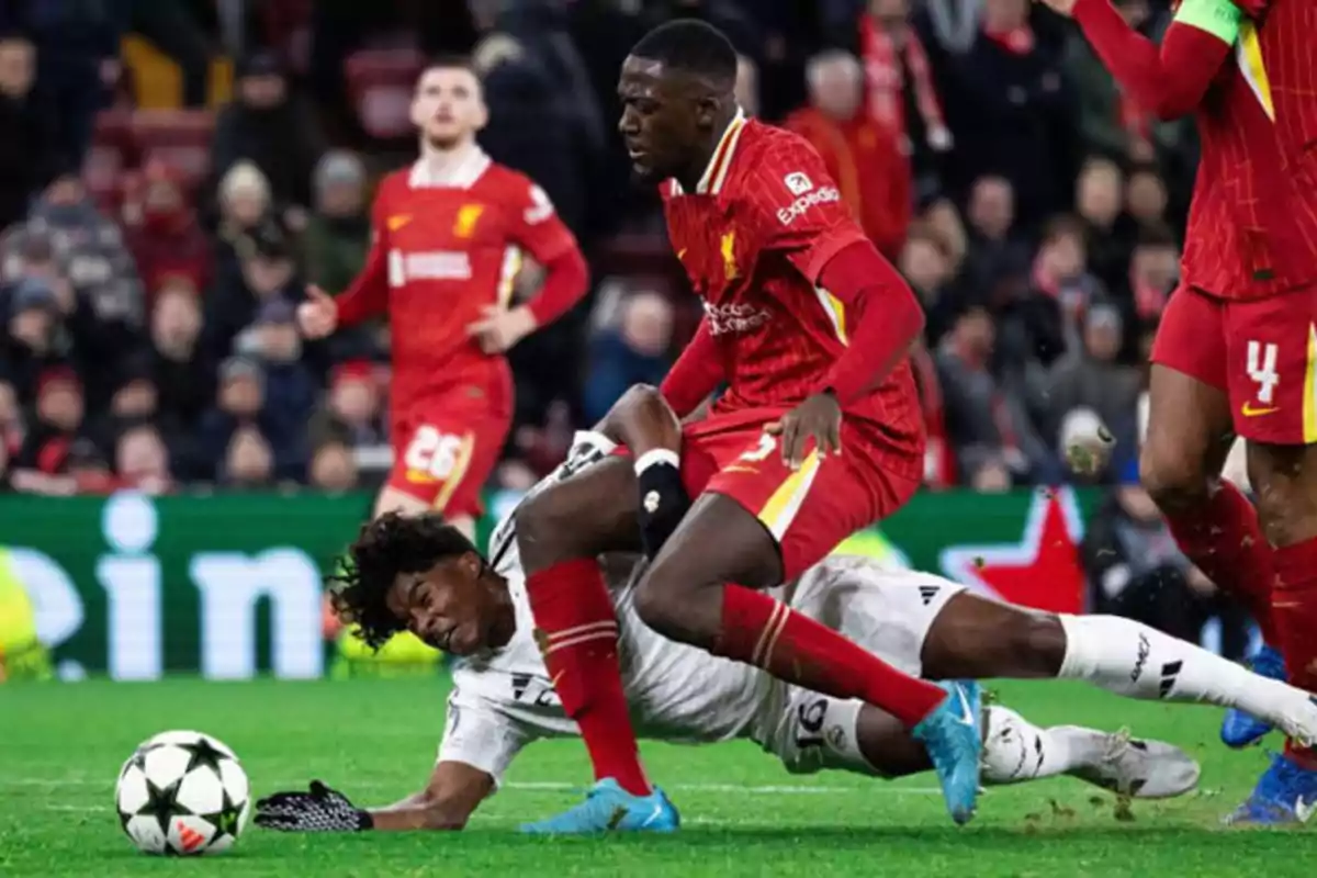 Football players in action during a match, one of them falls to the ground while the other tries to recover the ball.