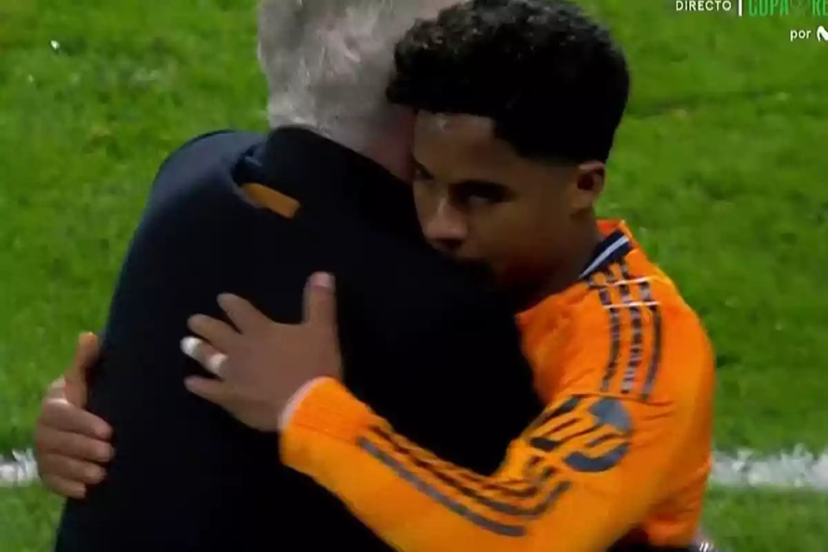 Dos personas se abrazan en un campo de fútbol, una de ellas lleva una chaqueta oscura y la otra un uniforme deportivo naranja. Dos personas se abrazan en un campo de fútbol, una de ellas lleva una chaqueta oscura y la otra un uniforme deportivo naranja.