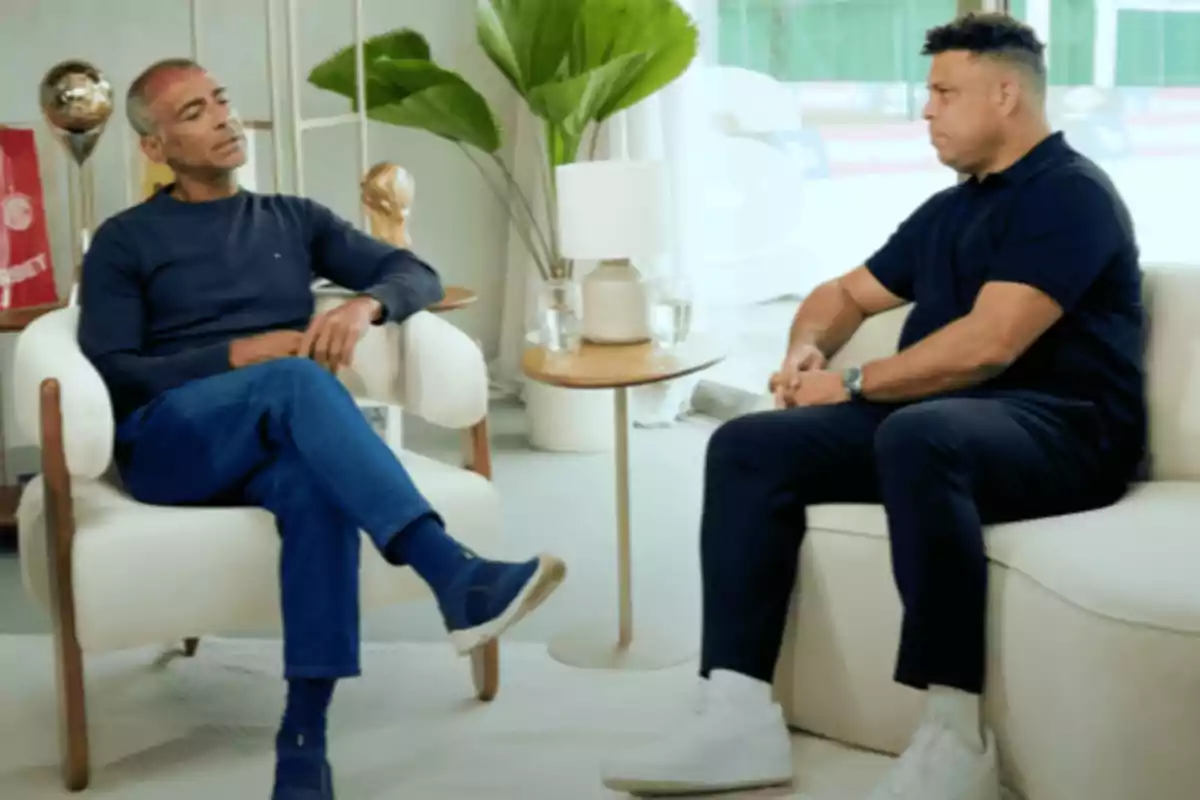 Two people sitting in white armchairs are conversing in a modern room decorated with plants and trophies.