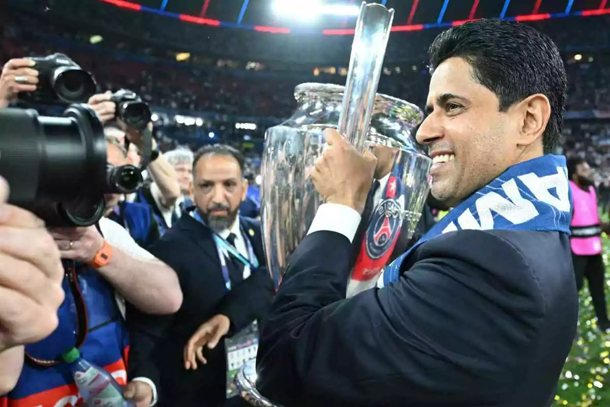 Smiling man with a blue scarf holds a silver trophy while photographers surround him in a stadium full of spectators Smiling man with a blue scarf holds a silver trophy while photographers surround him in a stadium full of spectators