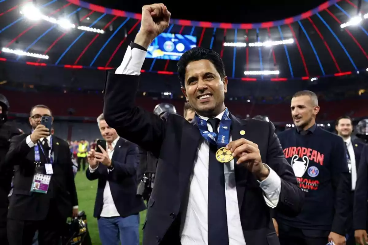 Hombre sonriente con medalla de campeón levantando el brazo en señal de victoria en un estadio iluminado rodeado de personas celebrando y tomando fotos