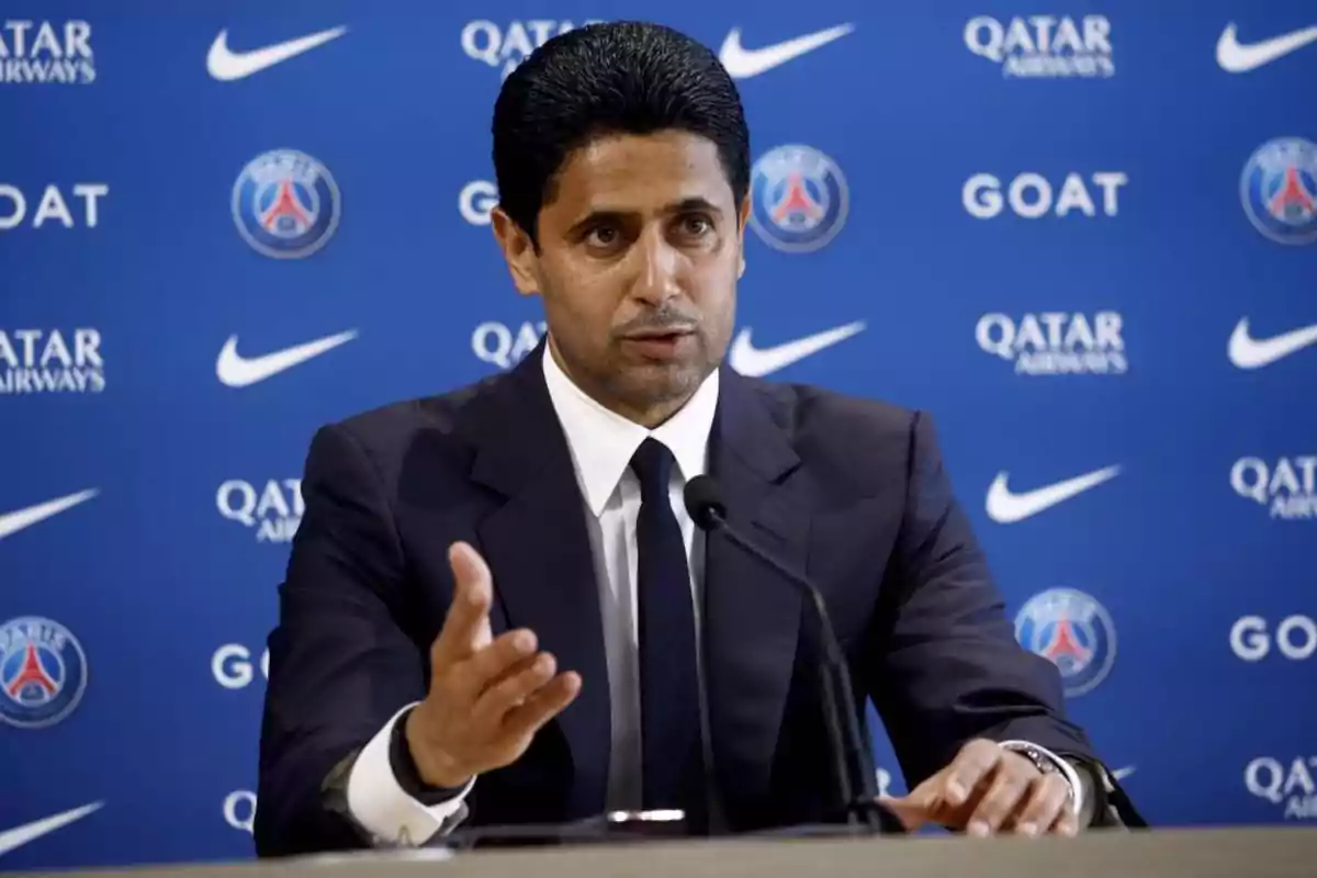 A man in a suit speaking at a press conference against a blue background displaying logos of Qatar Airways, Nike and PSG. A man in a suit speaking at a press conference against a blue background displaying logos of Qatar Airways, Nike and PSG.