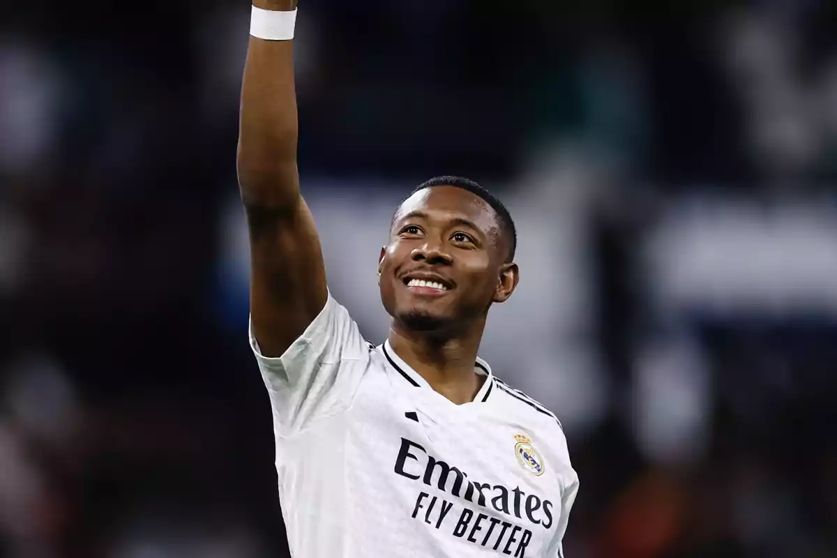 Un jugador de fútbol con uniforme blanco levanta el brazo y sonríe en un estadio.