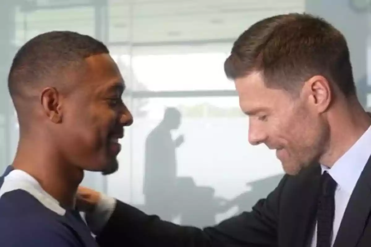 Two men greet each other with smiles in an indoor setting with a blurred background.