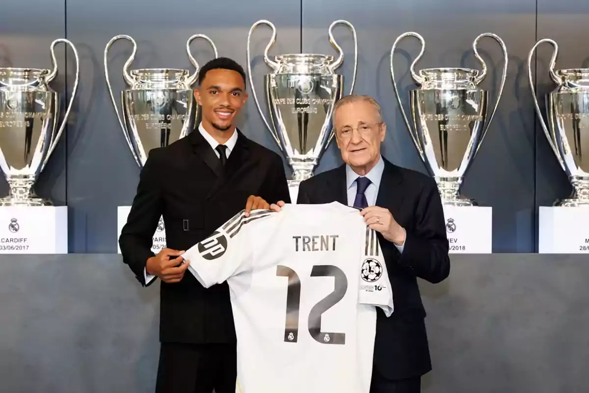 Two men pose smiling, holding a white jersey with the name Trent and the number 12 in front of several Champions League trophies. Two men pose smiling, holding a white jersey with the name Trent and the number 12 in front of several Champions League trophies.
