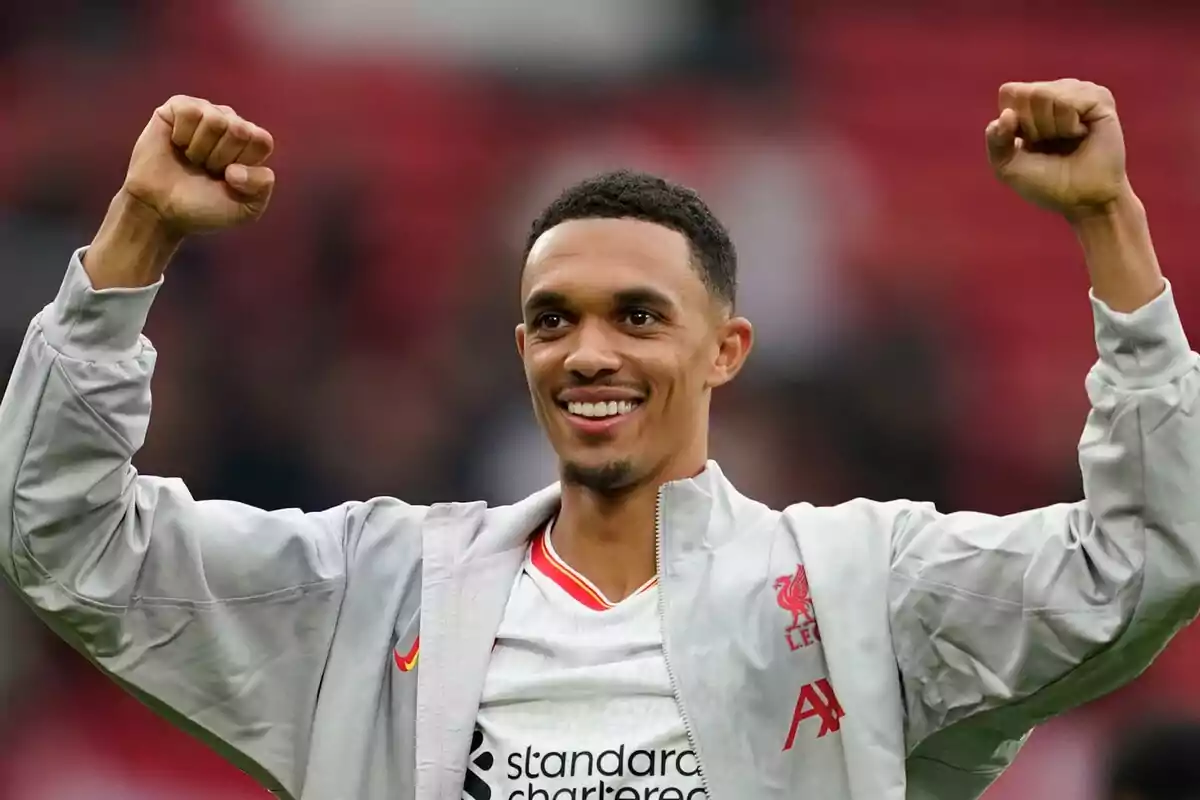 A soccer player smiles and raises his arms in victory, wearing a grey jacket and a Liverpool jersey. A soccer player smiles and raises his arms in victory, wearing a grey jacket and a Liverpool jersey.